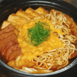 Fully cooked budae jjigae topped with ramen noodles, melted American cheese, and scallion curls in a black pot.