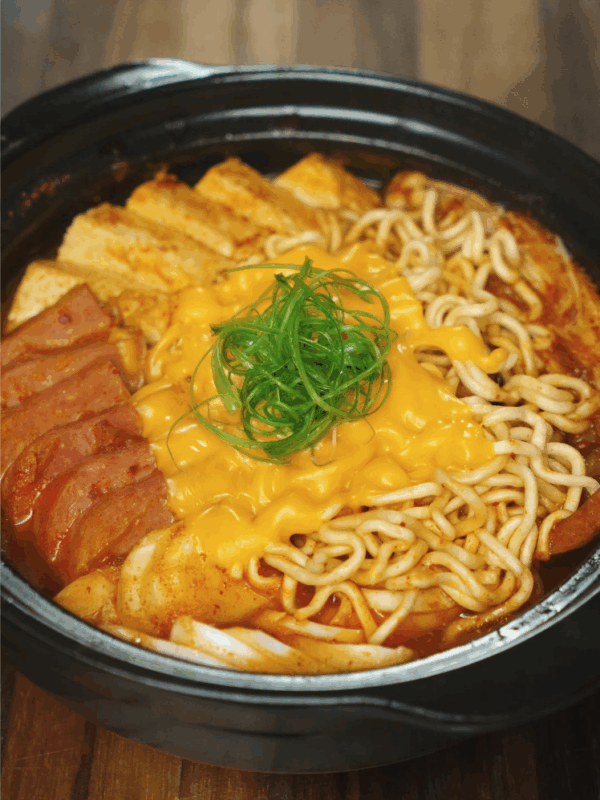 Fully cooked budae jjigae topped with ramen noodles, melted American cheese, and scallion curls in a black pot.