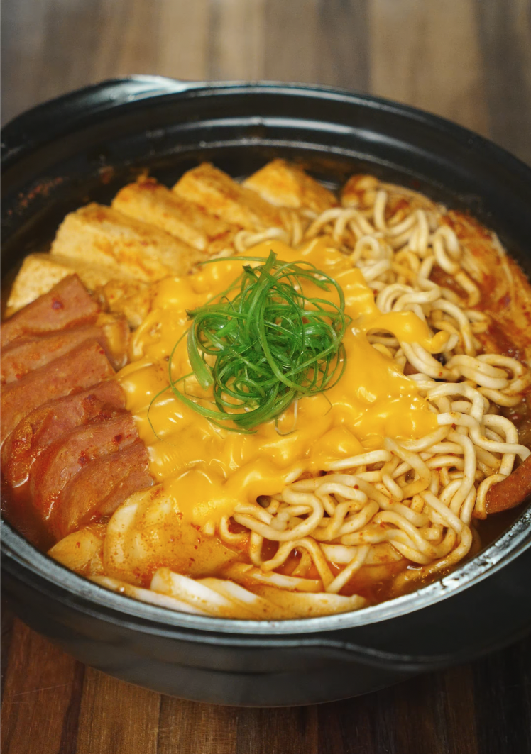 Fully cooked budae jjigae topped with ramen noodles, melted American cheese, and scallion curls in a black pot.