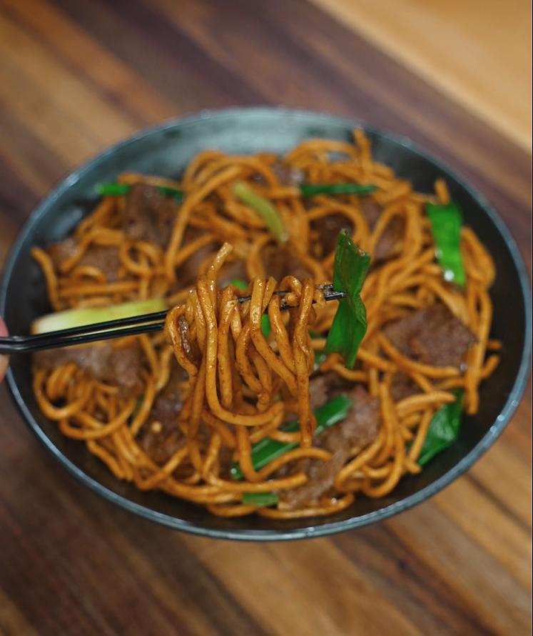 A close-up of chopsticks lifting a bite of beef lo mein, showcasing glossy noodles wrapped around stir-fried green onions and tender beef. This action shot highlights the texture and savory appeal of the dish.