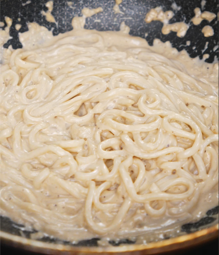 Freshly cooked noodles being stirred into the creamy garlic sauce, evenly coated and thickening in the pan.