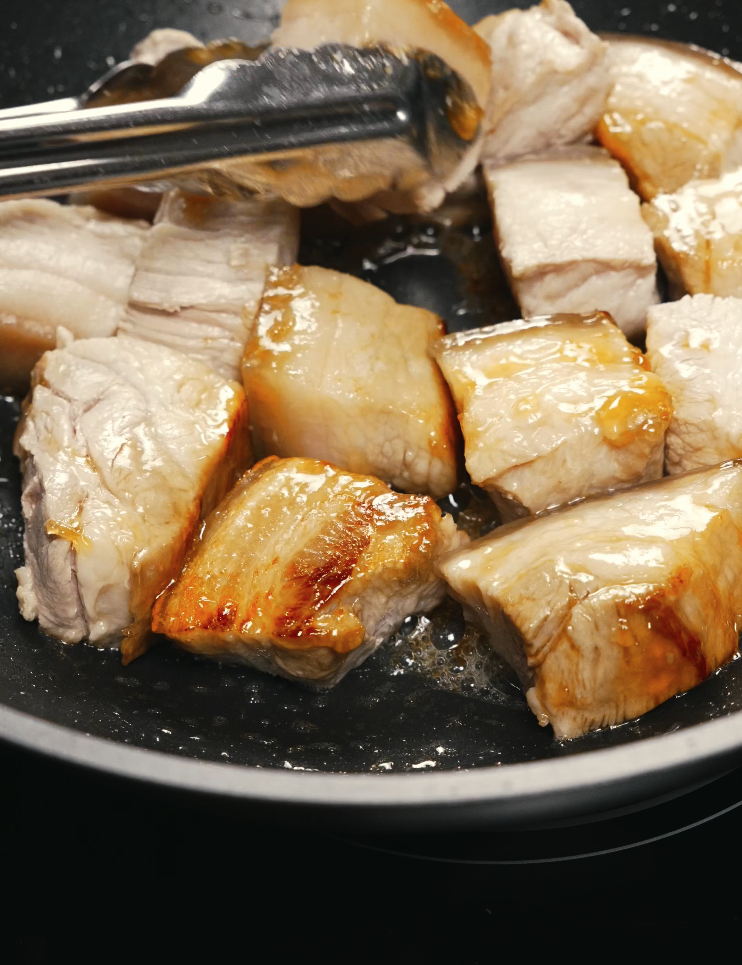 Pork belly cubes being pan-seared with tongs in a skillet, browning the exterior to add flavor before braising.