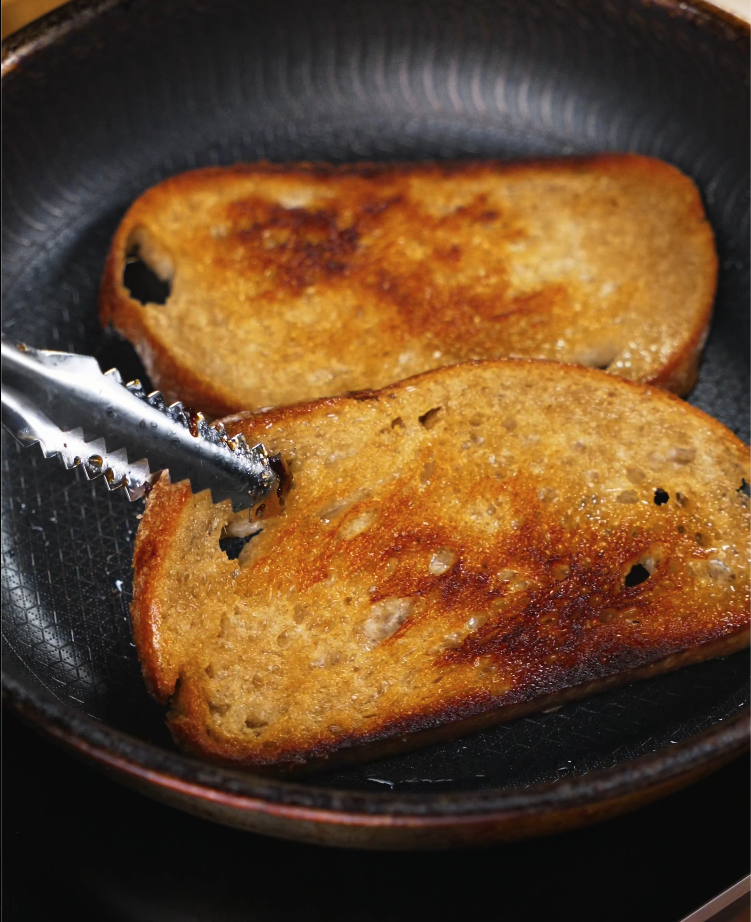 Two slices of sourdough bread being toasted in a nonstick pan until golden brown and crisp.