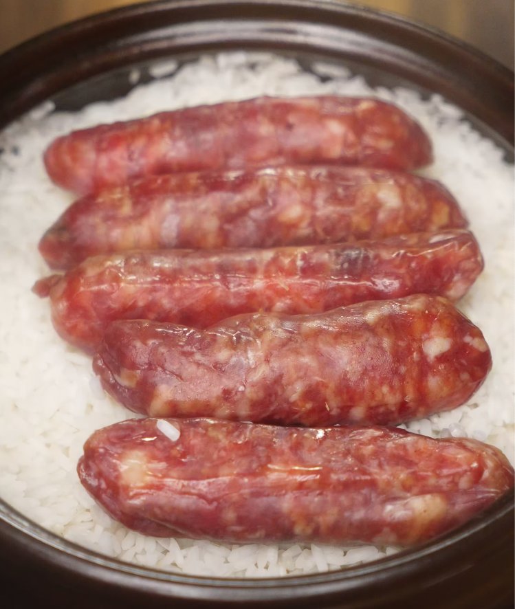Five raw Chinese sausages arranged in a single layer on top of uncooked rice in a claypot, preparing to be cooked together for flavor infusion.