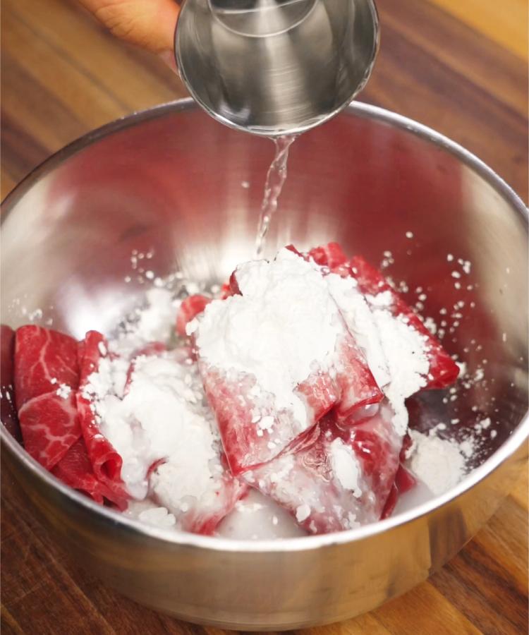 Beef slices being evenly coated in a cornstarch slurry inside a metal mixing bowl, preparing the meat for quick stir-frying. This method enhances the texture and ensures the beef stays tender in the final Beef Lo Mein dish.