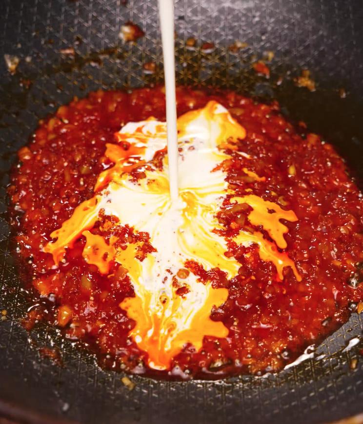 Heavy cream being poured into a bubbling mixture of gochujang and aromatics in a pan, beginning the process of creating a rich and spicy Alfredo sauce.