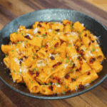 A full bowl of creamy gochujang rigatoni garnished with chili oil, parmesan, and green herbs, served in a dark ceramic dish on a wooden surface.