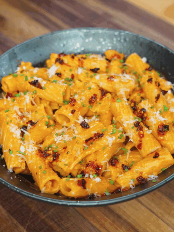 A full bowl of creamy gochujang rigatoni garnished with chili oil, parmesan, and green herbs, served in a dark ceramic dish on a wooden surface.