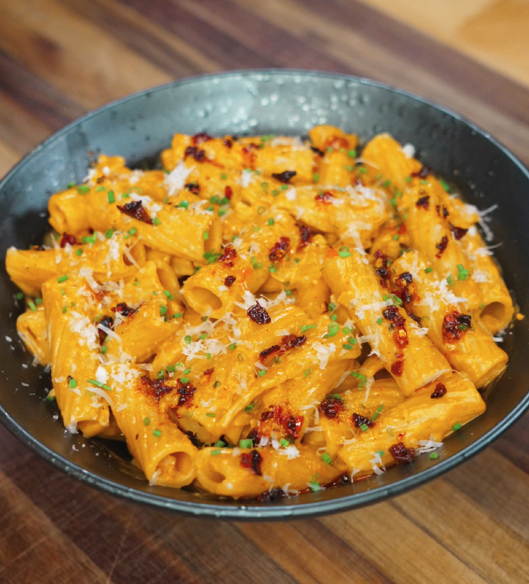 A full bowl of creamy gochujang rigatoni garnished with chili oil, parmesan, and green herbs, served in a dark ceramic dish on a wooden surface.