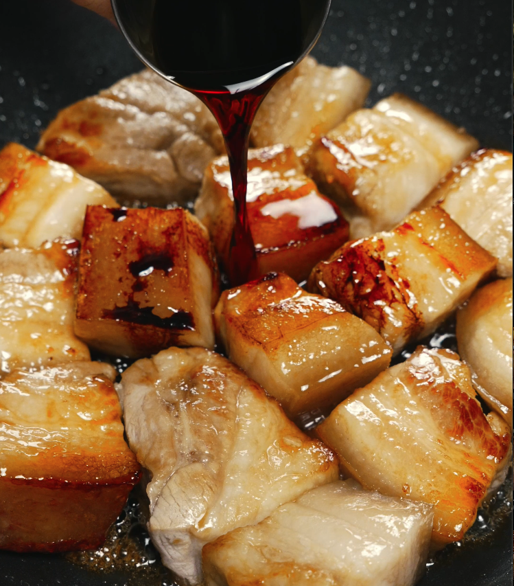 Dark soy sauce being poured onto seared pork belly pieces in a pan, setting the foundation for the rich braising sauce.