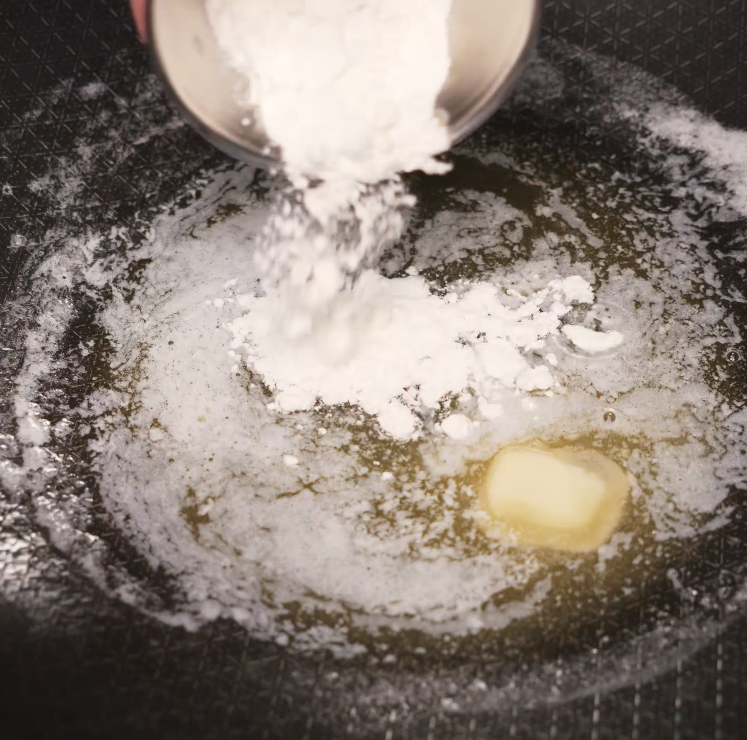 Flour is being poured into a pan of melted butter to create a roux base, which will serve as the foundation for the creamy gochujang cheese sauce.