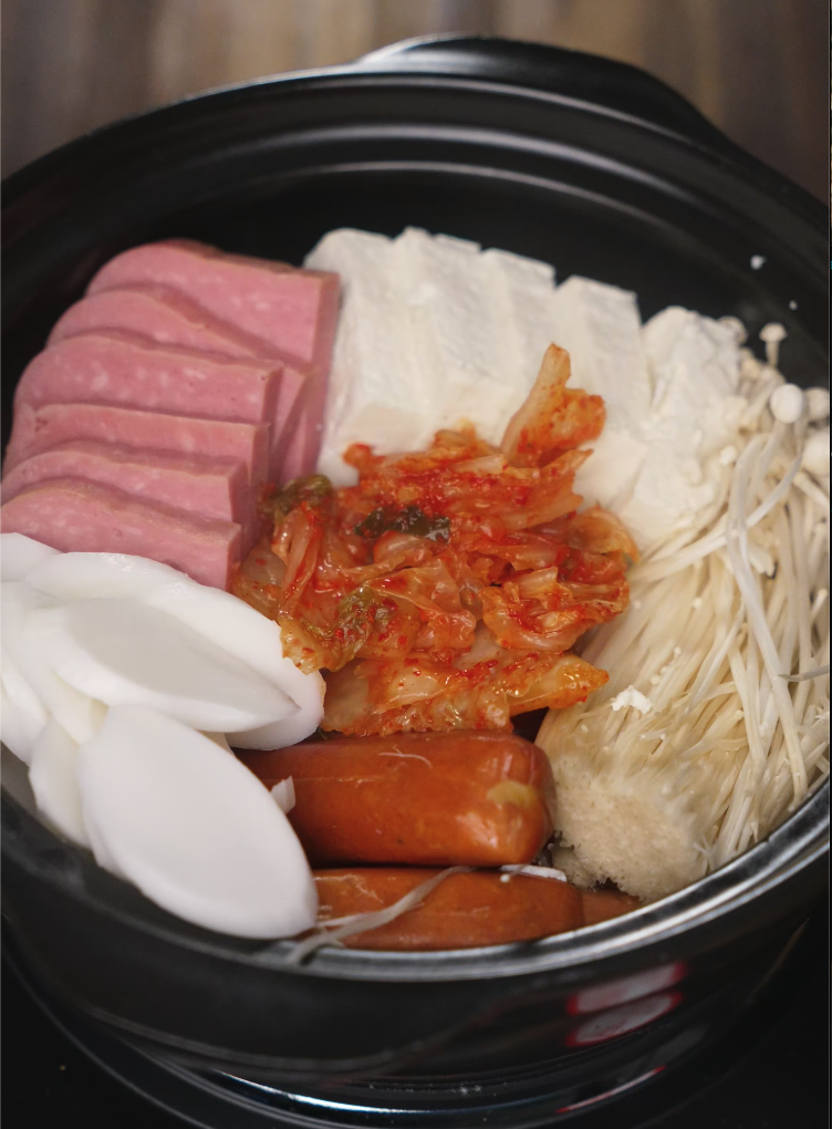 Arranging ingredients like sliced spam, tofu, kimchi, rice cakes, and mushrooms into a shallow Korean hot pot.