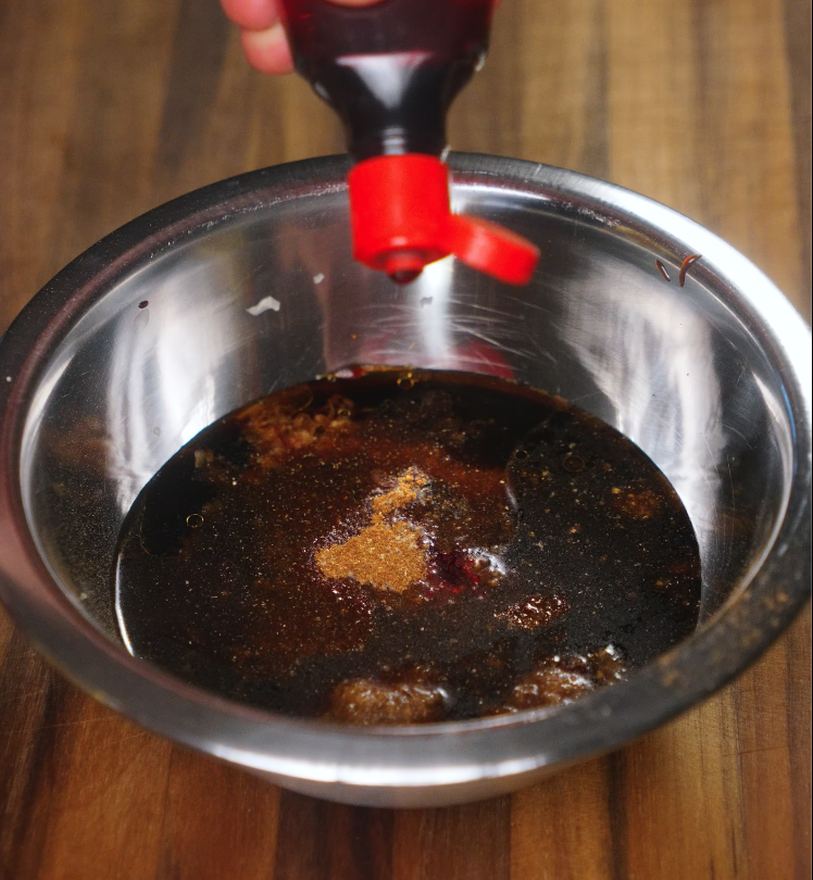 A hand pouring hoisin and soy-based marinade into a metal bowl with brown sugar and Chinese five spice for char siu pork preparation.