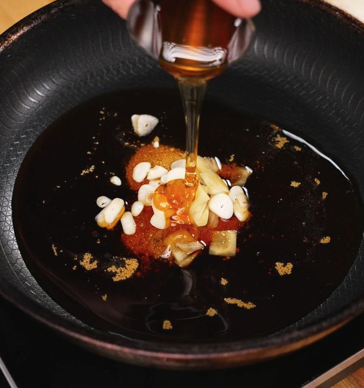 A glass container of honey is being poured into a pan containing garlic, ginger, and soy sauce, forming the rich and aromatic braising base for the chicken.