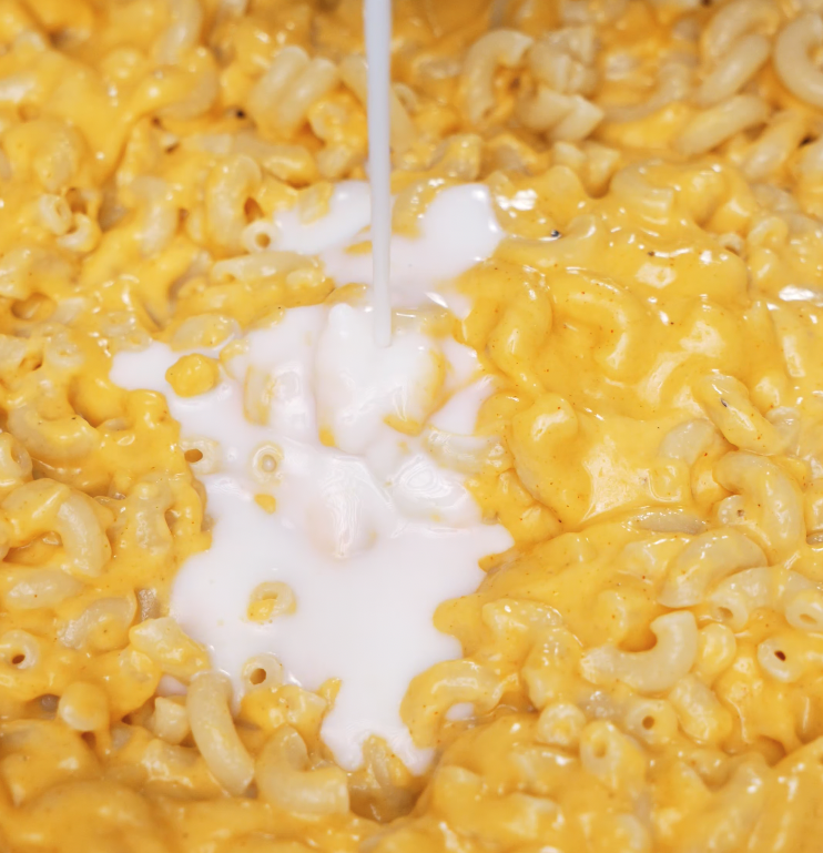 Milk is poured into a pan of roux and gochujang, forming a smooth, vibrant orange cheese sauce base for gochujang mac and cheese.