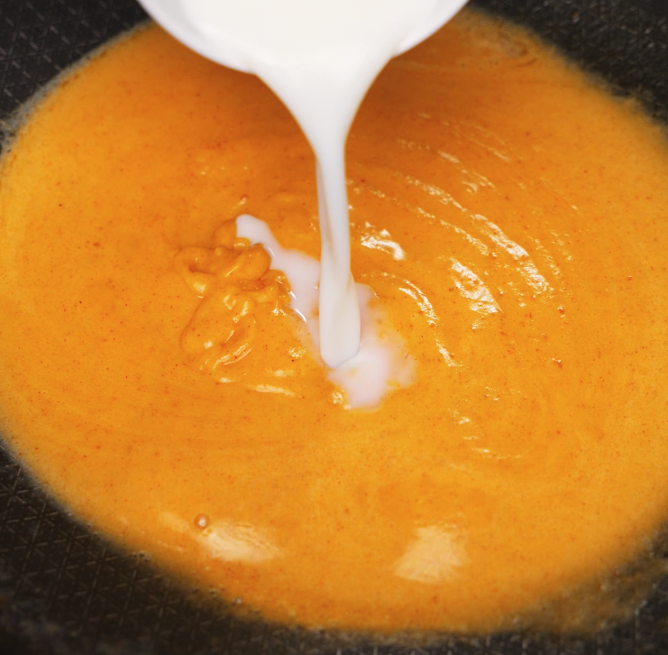 Milk is poured into a pan of roux and gochujang, forming a smooth, vibrant orange cheese sauce base for gochujang mac and cheese.
