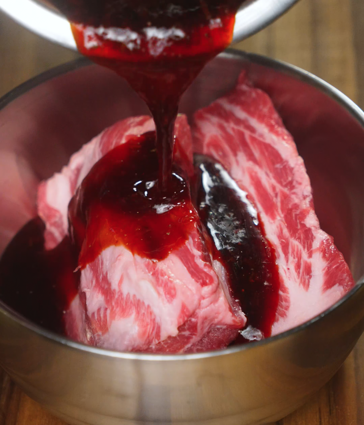 Thick red marinade being poured over pork shoulder pieces in a mixing bowl for Chinese char siu preparation.