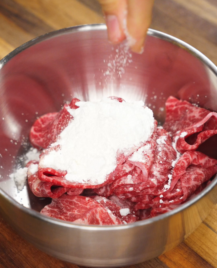 Raw thinly sliced beef being dusted with cornstarch in a metal mixing bowl, prepping the meat for stir-frying in the yaki udon recipe.