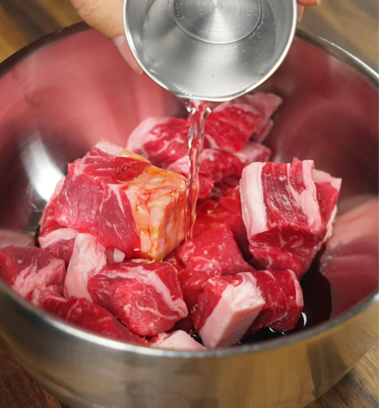 A close-up of water being poured from a metal measuring cup into a stainless steel bowl of cubed beef short ribs with marinade, prepping for a flavorful steak fried rice base.