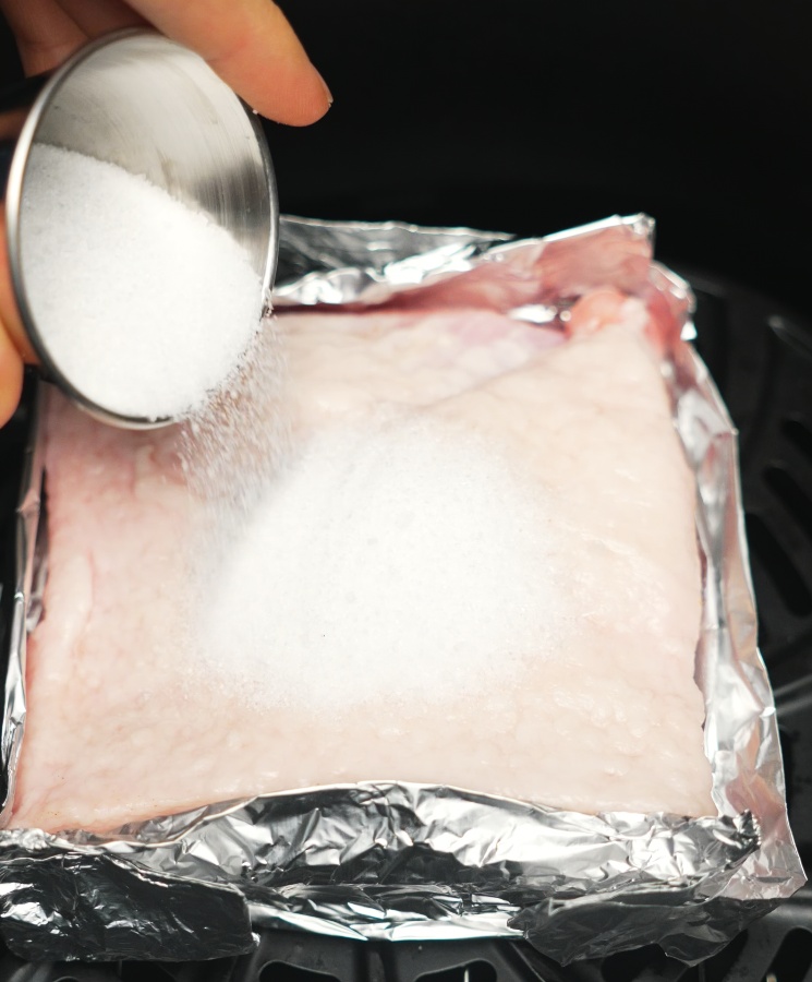 A layer of salt being evenly poured on top of the pork belly skin placed in a foil-lined tray to dry out the skin before air frying.
