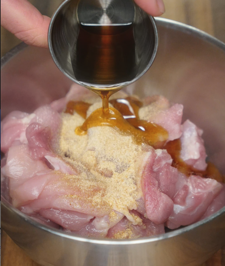 Pouring marinade over raw chicken in a metal bowl. The marinade includes soy sauce, garlic powder, and other seasonings.
