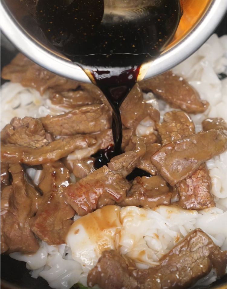 Pouring a thick soy sauce-based mixture over tender beef slices and rice noodles in the wok to begin building the glossy, flavorful glaze for Beef Chow Fun. This step ensures even distribution of flavor.