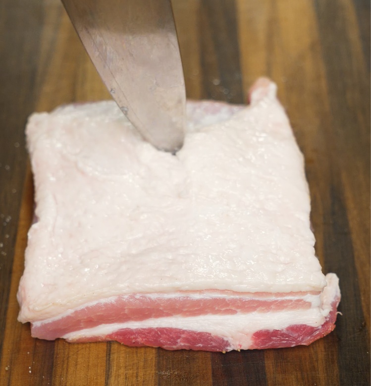 A knife scoring the top fat layer of raw pork belly to help render fat and ensure an even crispy texture after roasting.
