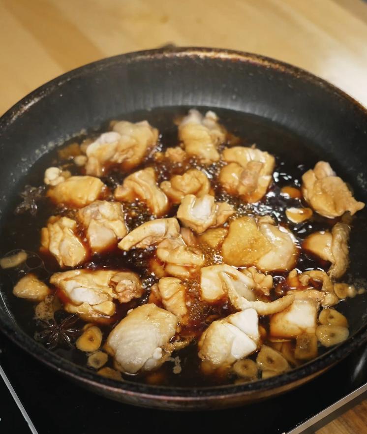 Chunks of chicken thigh are shown simmering in a dark soy-based sauce with visible slices of ginger and garlic, absorbing deep umami flavors.