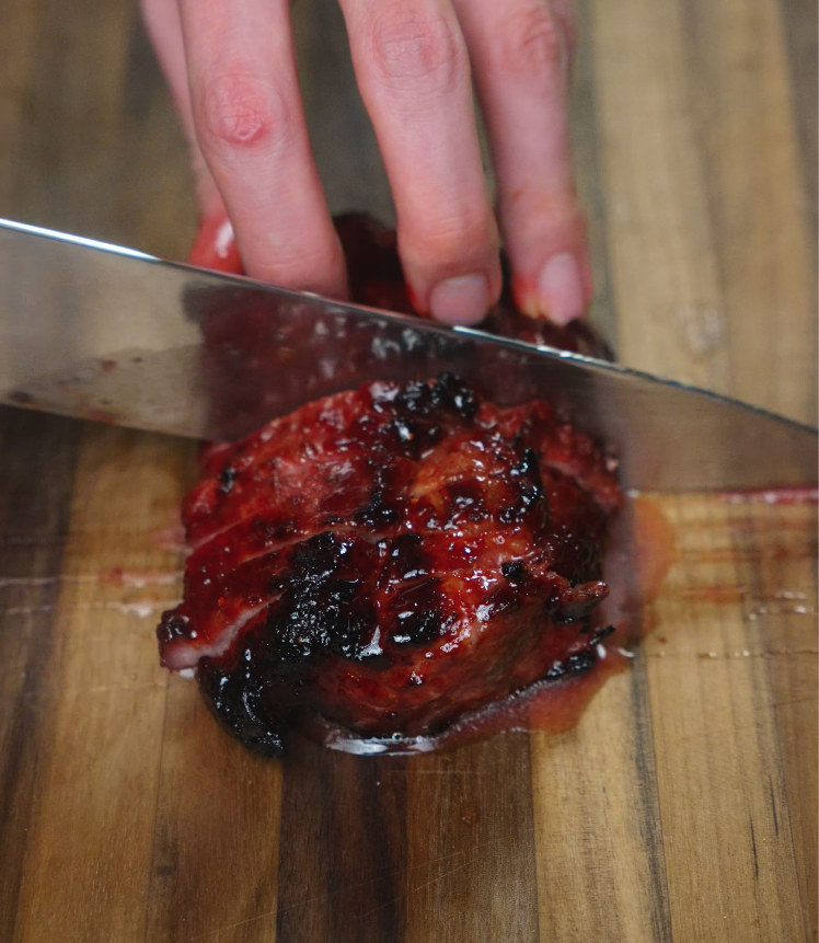 Hand slicing through cooked char siu pork on a wooden cutting board, showing the tender and glossy interior texture.