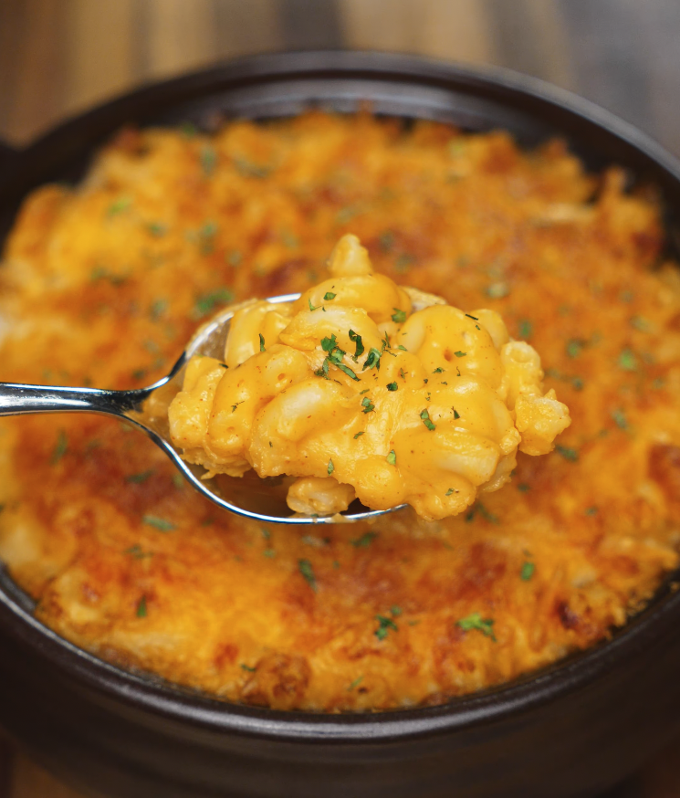 A spoonful of gochujang mac and cheese is lifted above the baked dish, showcasing the creamy texture and golden cheese topping.