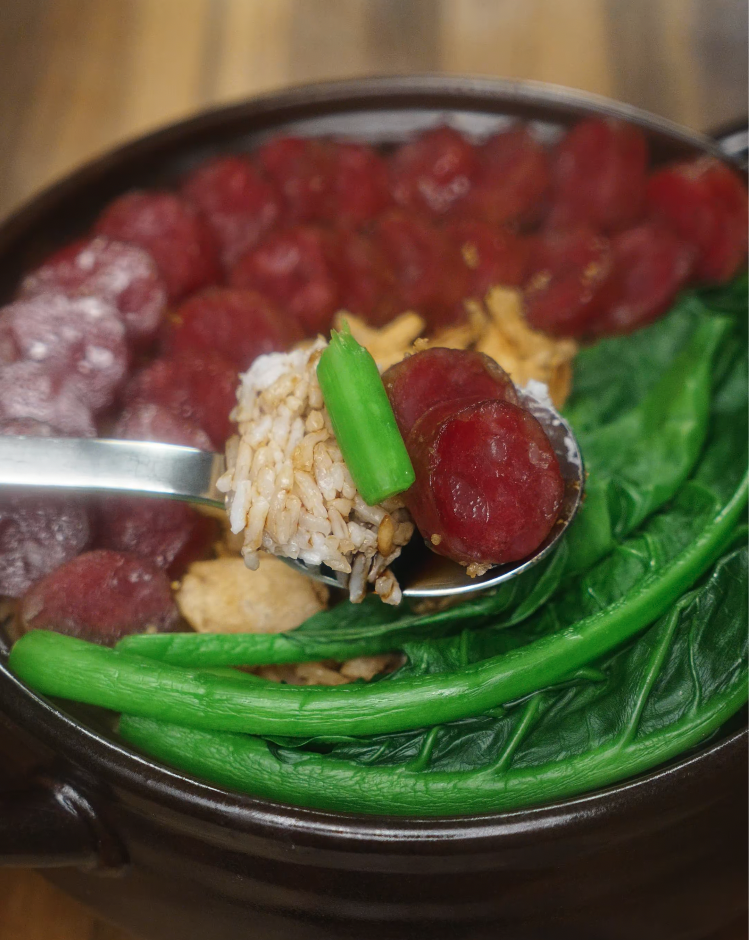 A spoonful of finished claypot rice lifted from the pot, showcasing a bite with Chinese sausage, green choy sum, and soy-flavored rice.