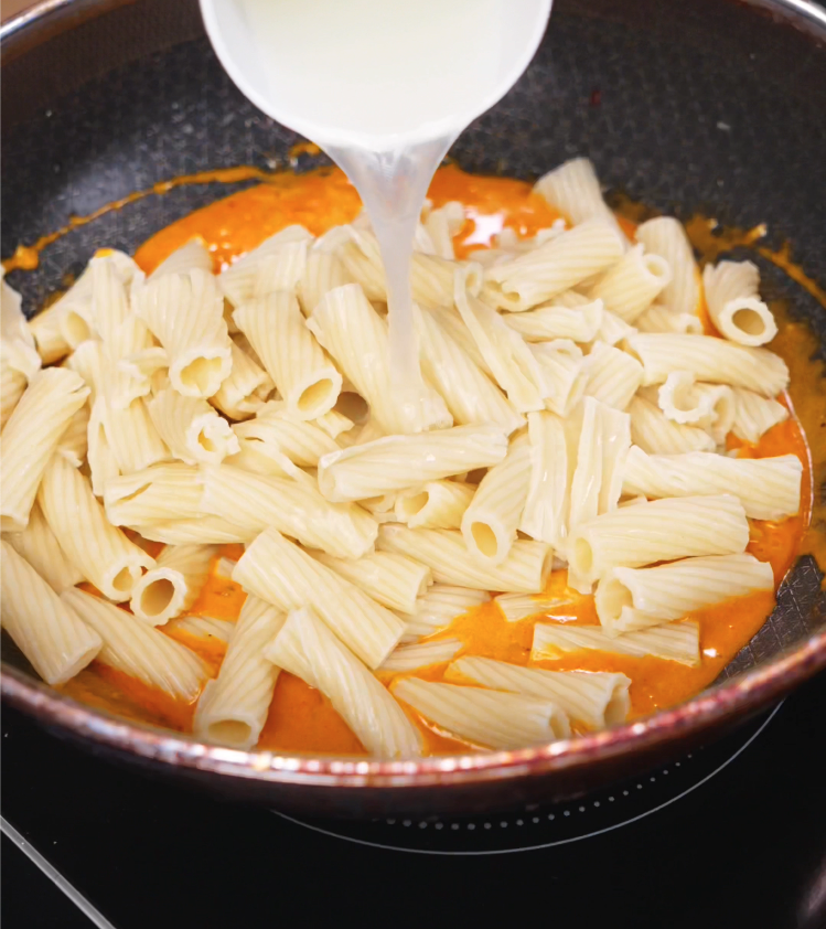 Cooked rigatoni and a splash of reserved pasta water being added to the creamy gochujang sauce in a pan, just before tossing everything together.