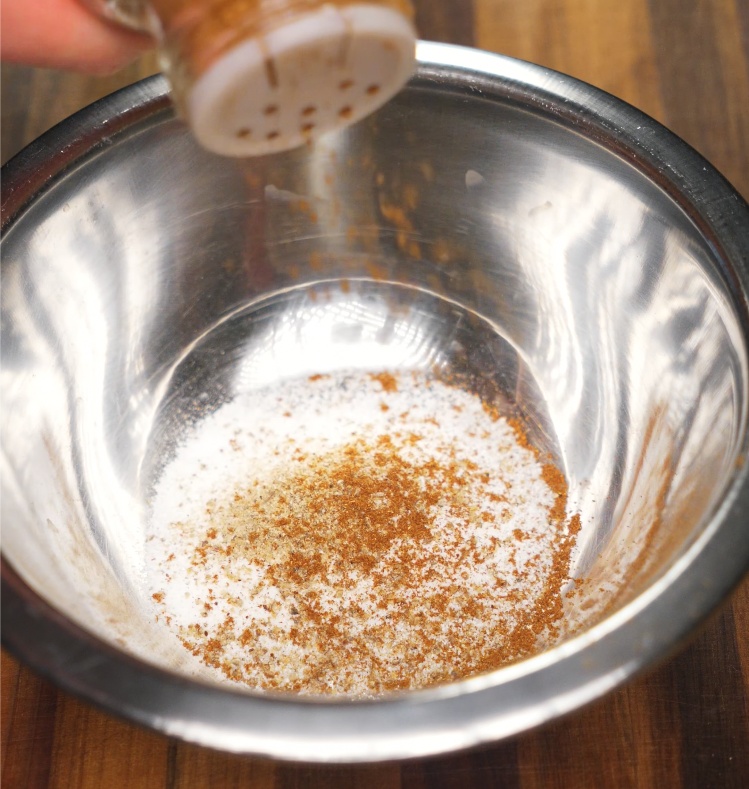Ground five spice powder and salt being sprinkled into a stainless steel mixing bowl, resting on a wooden kitchen counter to prepare the pork belly seasoning.