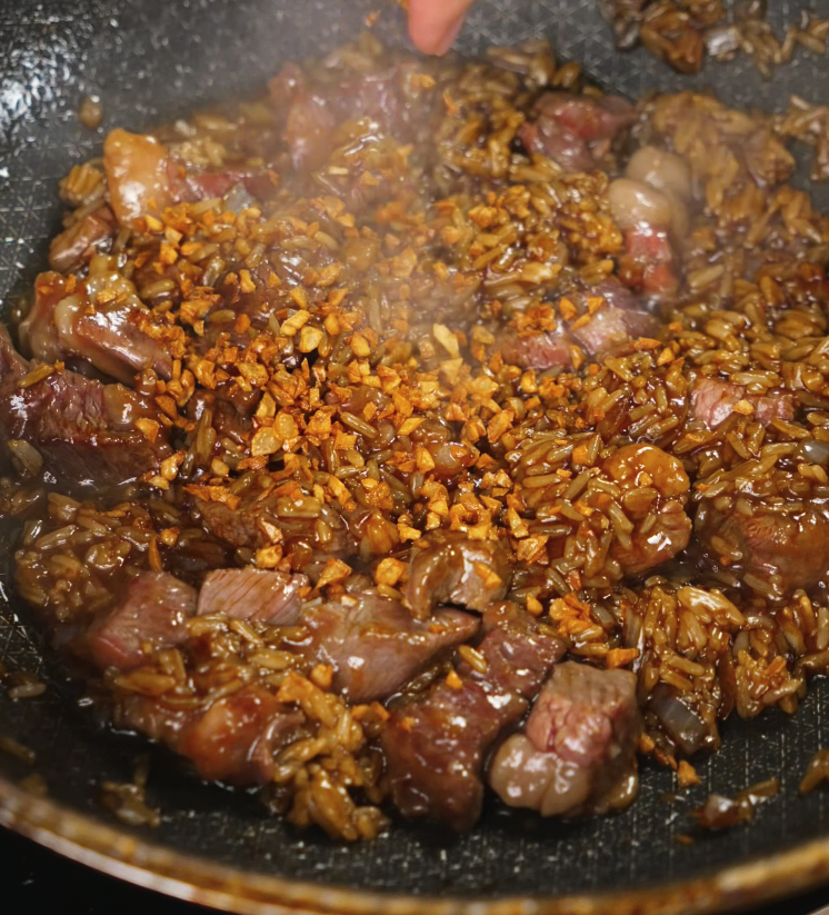 A frying pan filled with seared steak cubes and cooked rice being tossed together with crispy garlic for a rich, savory garlic steak fried rice.