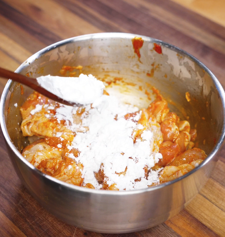 A spoonful of white cornstarch is being added to a metal bowl of chicken thighs already marinated in a reddish-orange spice blend. The cornstarch will help create a light, crispy coating when the chicken is air-fried.