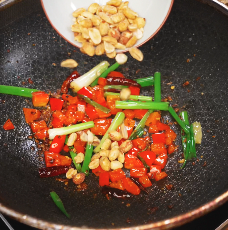 A bowl of roasted peanuts is tipped over a pan filled with stir-fried red bell peppers, scallions, and dried chilies. These ingredients form the signature crunchy, nutty texture and colorful base of kung pao chicken.