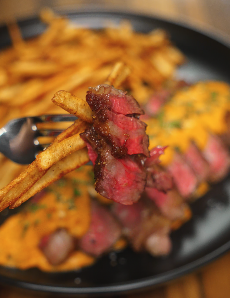 A fork lifting a bite of juicy ribeye and a few five-spice fries above a plate of steak and sauce.