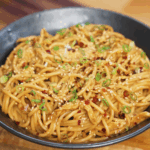 Close-up of creamy chicken peanut butter noodles served in a dark bowl, topped generously with chopped green onions, toasted sesame seeds, and red pepper flakes. The glossy peanut sauce coats every strand of spaghetti, creating a rich and flavorful finish perfect for an easy Asian-inspired dinner.