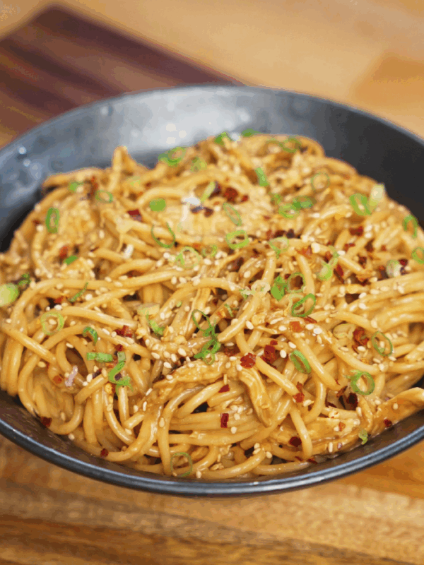 Close-up of creamy chicken peanut butter noodles served in a dark bowl, topped generously with chopped green onions, toasted sesame seeds, and red pepper flakes. The glossy peanut sauce coats every strand of spaghetti, creating a rich and flavorful finish perfect for an easy Asian-inspired dinner.