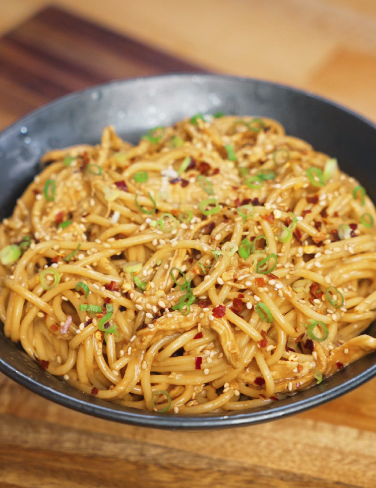 Close-up of creamy chicken peanut butter noodles served in a dark bowl, topped generously with chopped green onions, toasted sesame seeds, and red pepper flakes. The glossy peanut sauce coats every strand of spaghetti, creating a rich and flavorful finish perfect for an easy Asian-inspired dinner.