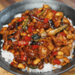 A bowl of kung pao chicken served over fluffy white rice, featuring tender chicken pieces, roasted peanuts, red bell peppers, scallions, and dried chili peppers all coated in a glossy, spicy-sweet sauce. The dish is vibrant, colorful, and packed with classic Sichuan flavor.
