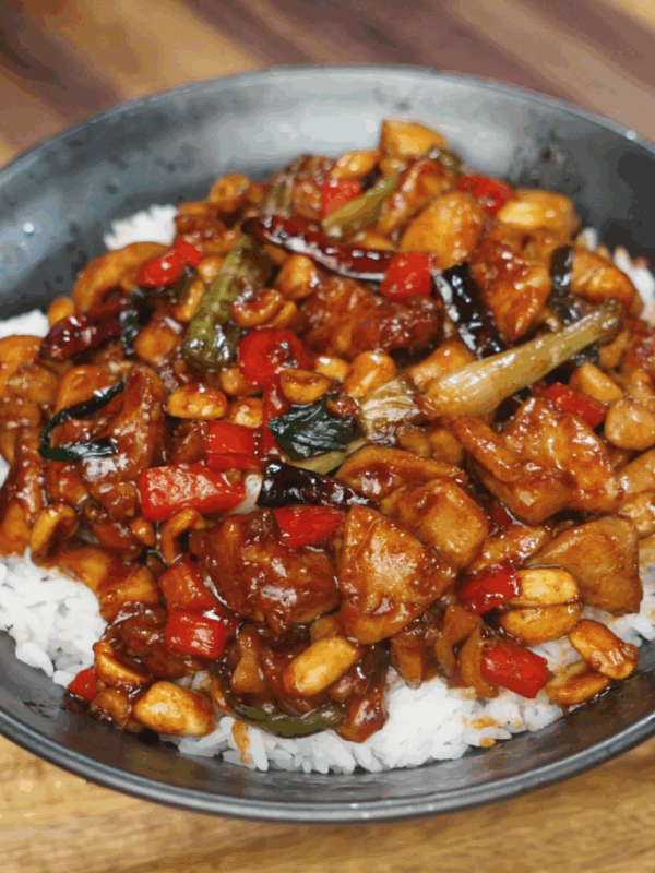 A bowl of kung pao chicken served over fluffy white rice, featuring tender chicken pieces, roasted peanuts, red bell peppers, scallions, and dried chili peppers all coated in a glossy, spicy-sweet sauce. The dish is vibrant, colorful, and packed with classic Sichuan flavor.