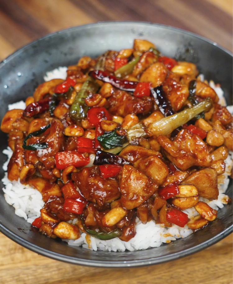 A bowl of kung pao chicken served over fluffy white rice, featuring tender chicken pieces, roasted peanuts, red bell peppers, scallions, and dried chili peppers all coated in a glossy, spicy-sweet sauce. The dish is vibrant, colorful, and packed with classic Sichuan flavor.