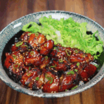 A beautifully plated garlic soy chicken bowl featuring sticky glazed chicken thighs arranged over white rice and topped with scallions and sesame seeds, served with a generous portion of shredded green lettuce on the side. The chicken is deeply caramelized with a rich glaze that contrasts the fresh greens and fluffy rice base.