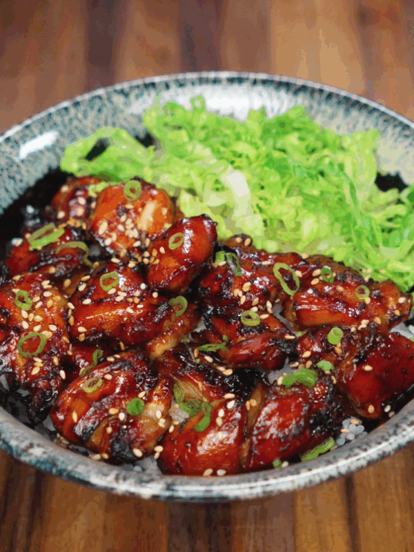 A beautifully plated garlic soy chicken bowl featuring sticky glazed chicken thighs arranged over white rice and topped with scallions and sesame seeds, served with a generous portion of shredded green lettuce on the side. The chicken is deeply caramelized with a rich glaze that contrasts the fresh greens and fluffy rice base.