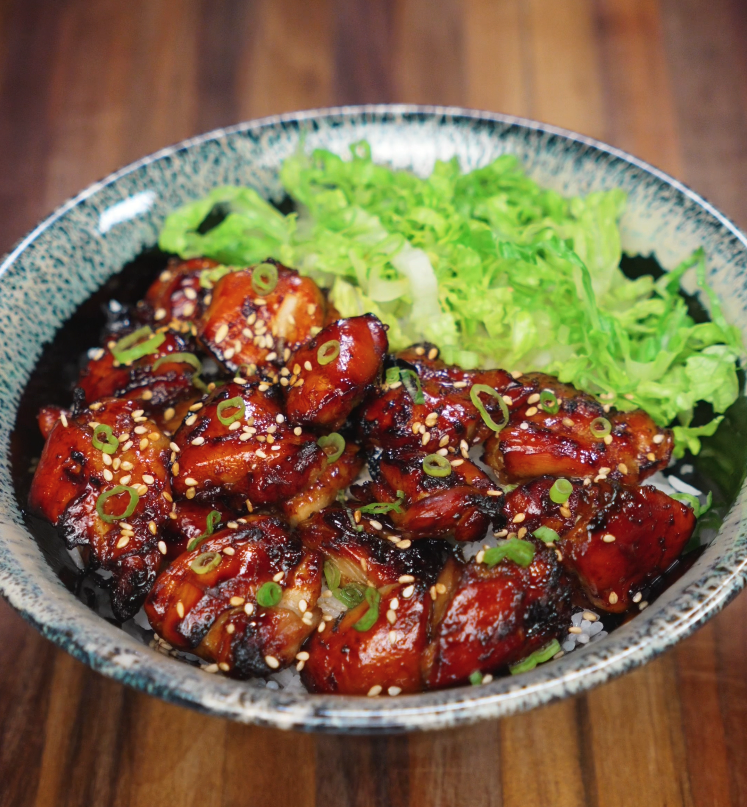 A beautifully plated garlic soy chicken bowl featuring sticky glazed chicken thighs arranged over white rice and topped with scallions and sesame seeds, served with a generous portion of shredded green lettuce on the side. The chicken is deeply caramelized with a rich glaze that contrasts the fresh greens and fluffy rice base.