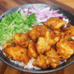 A bowl of golden crispy air-fried chicken thigh pieces served over fluffy white rice, accompanied by fresh shredded lettuce and thinly sliced red onions. The chicken is deeply caramelized, with a crunchy exterior and juicy center, making this a vibrant and satisfying rice bowl.