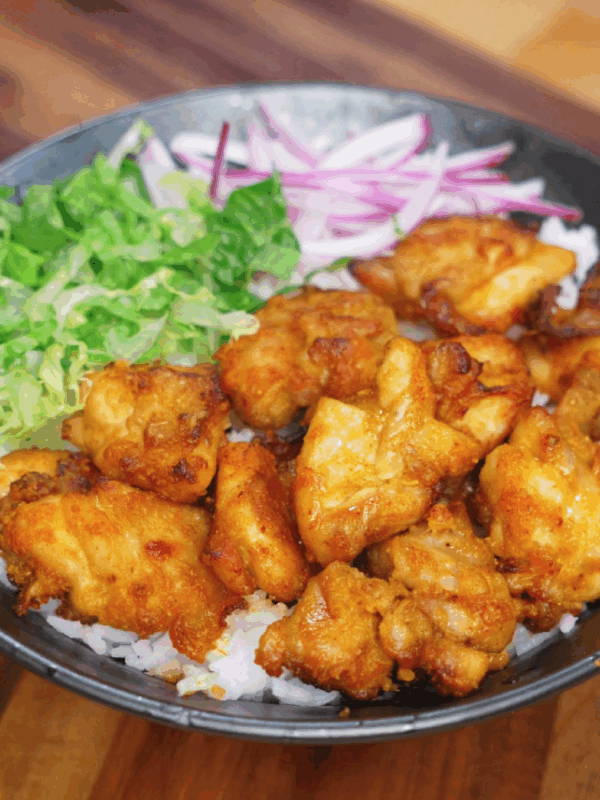 A bowl of golden crispy air-fried chicken thigh pieces served over fluffy white rice, accompanied by fresh shredded lettuce and thinly sliced red onions. The chicken is deeply caramelized, with a crunchy exterior and juicy center, making this a vibrant and satisfying rice bowl.