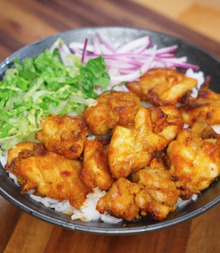 A bowl of golden crispy air-fried chicken thigh pieces served over fluffy white rice, accompanied by fresh shredded lettuce and thinly sliced red onions. The chicken is deeply caramelized, with a crunchy exterior and juicy center, making this a vibrant and satisfying rice bowl.
