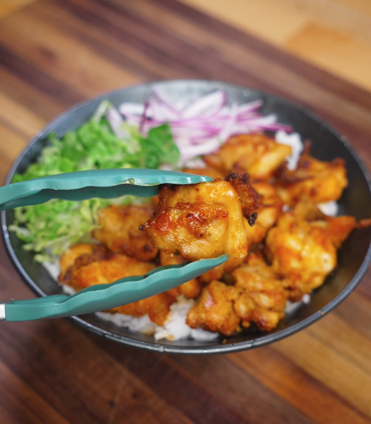 A juicy, golden-brown piece of air-fried chicken thigh is held up with teal tongs over a rice bowl layered with crispy chicken, fresh lettuce, and sliced red onions. The close-up highlights the crunchy coating and tender interior of the perfectly cooked chicken.